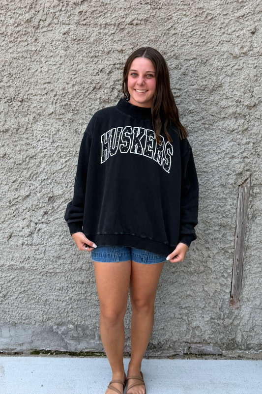 A woman stands against a textured concrete wall wearing a black oversized sweatshirt with 'HUSKERS' printed across the chest in white varsity-style lettering, paired with denim shorts and sandals.