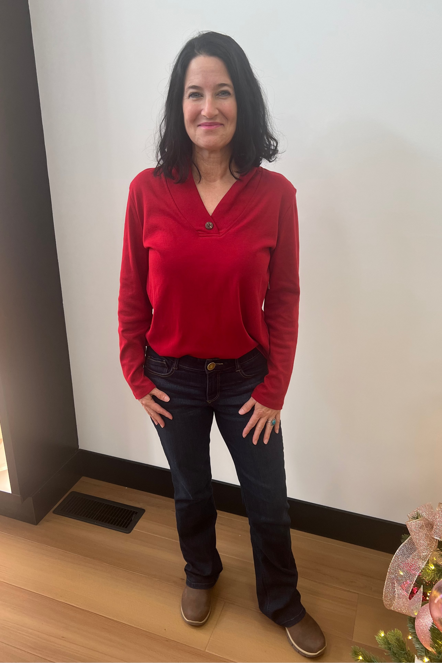 Woman wearing a red sweater and dark jeans standing indoors next to a Christmas tree.
