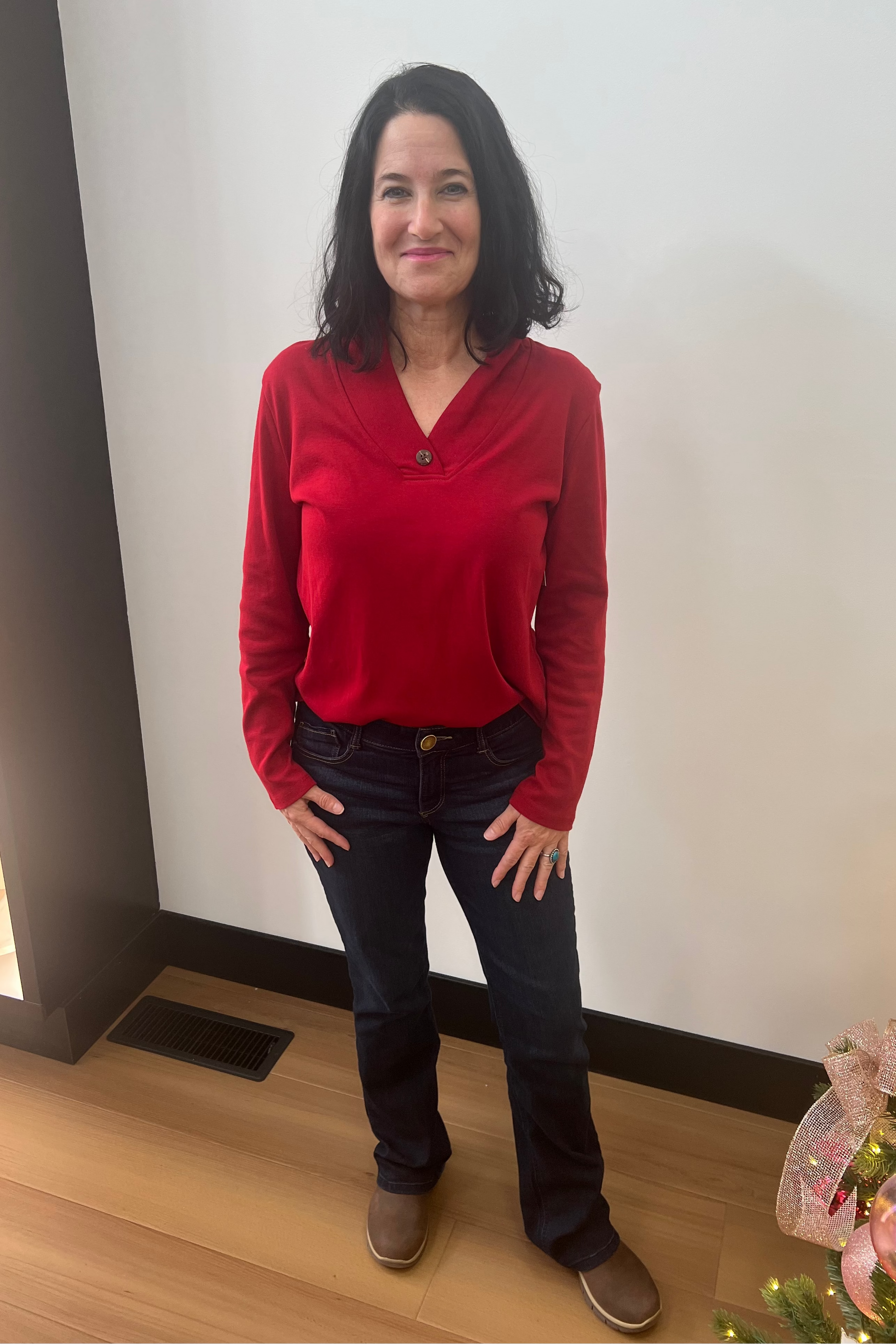 Woman wearing a red sweater and dark jeans standing indoors next to a Christmas tree.