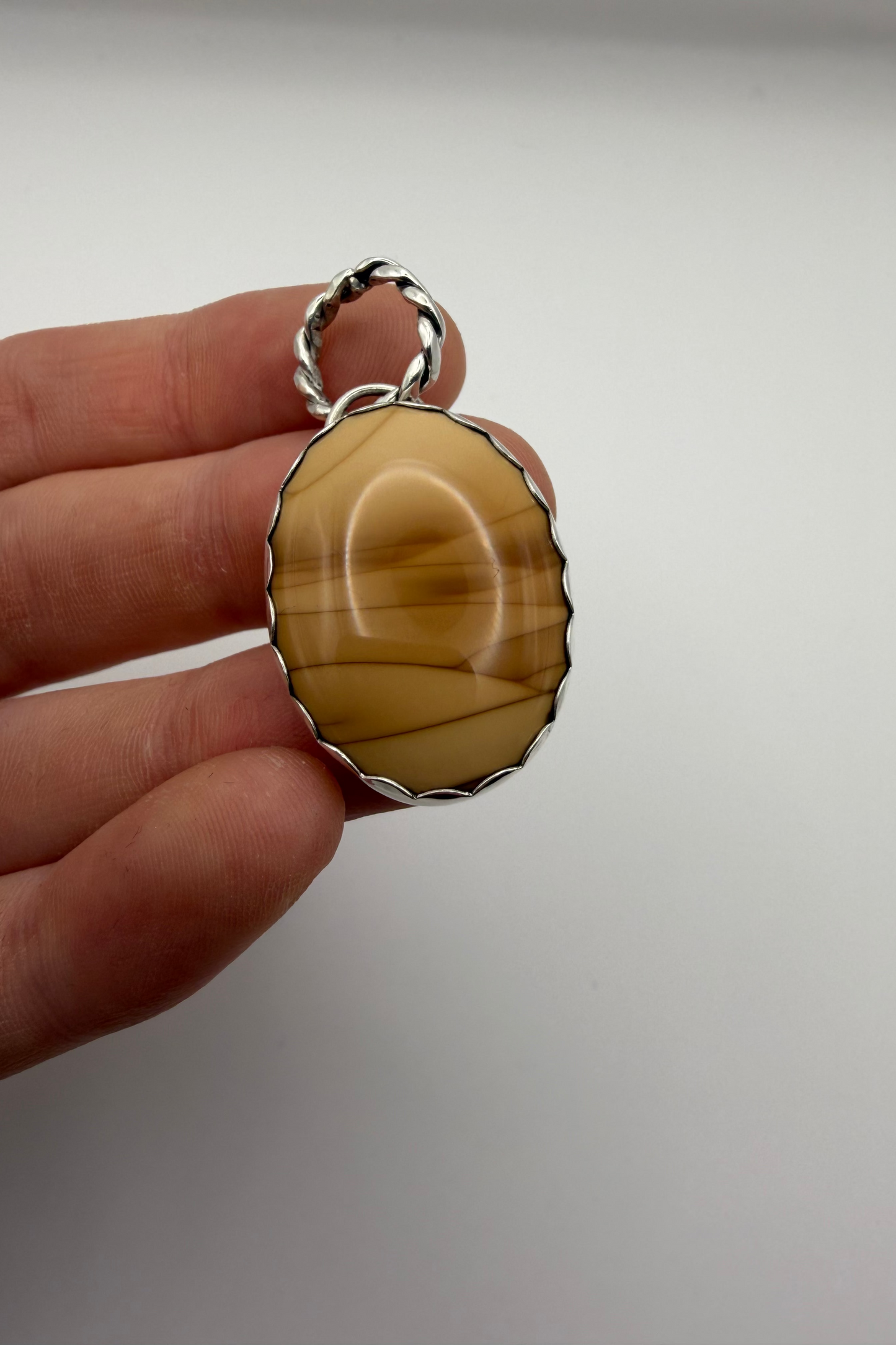 A hand holding a sterling silver pendant with a scalloped edge and a rope bail, featuring a smooth, polished oval stone with natural brown and cream banding.