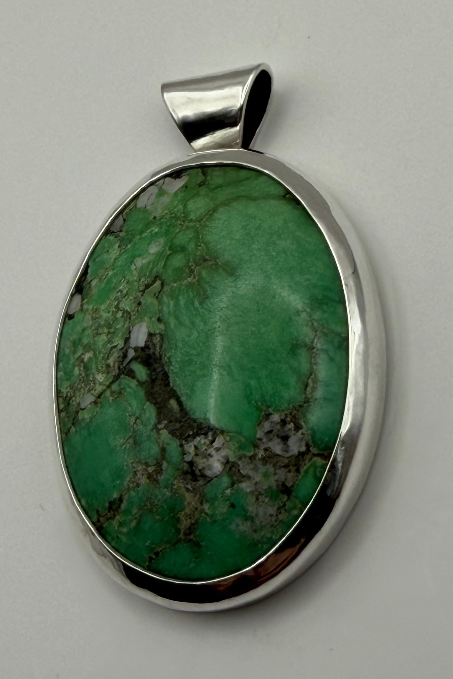 An oval pendant featuring a green stone with brown and white veining, set in a polished sterling silver bezel with a bail, shown on a white background.