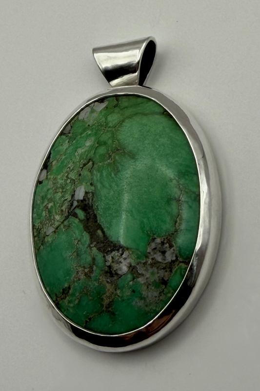 An oval pendant featuring a green stone with brown and white veining, set in a polished sterling silver bezel with a bail, shown on a white background.