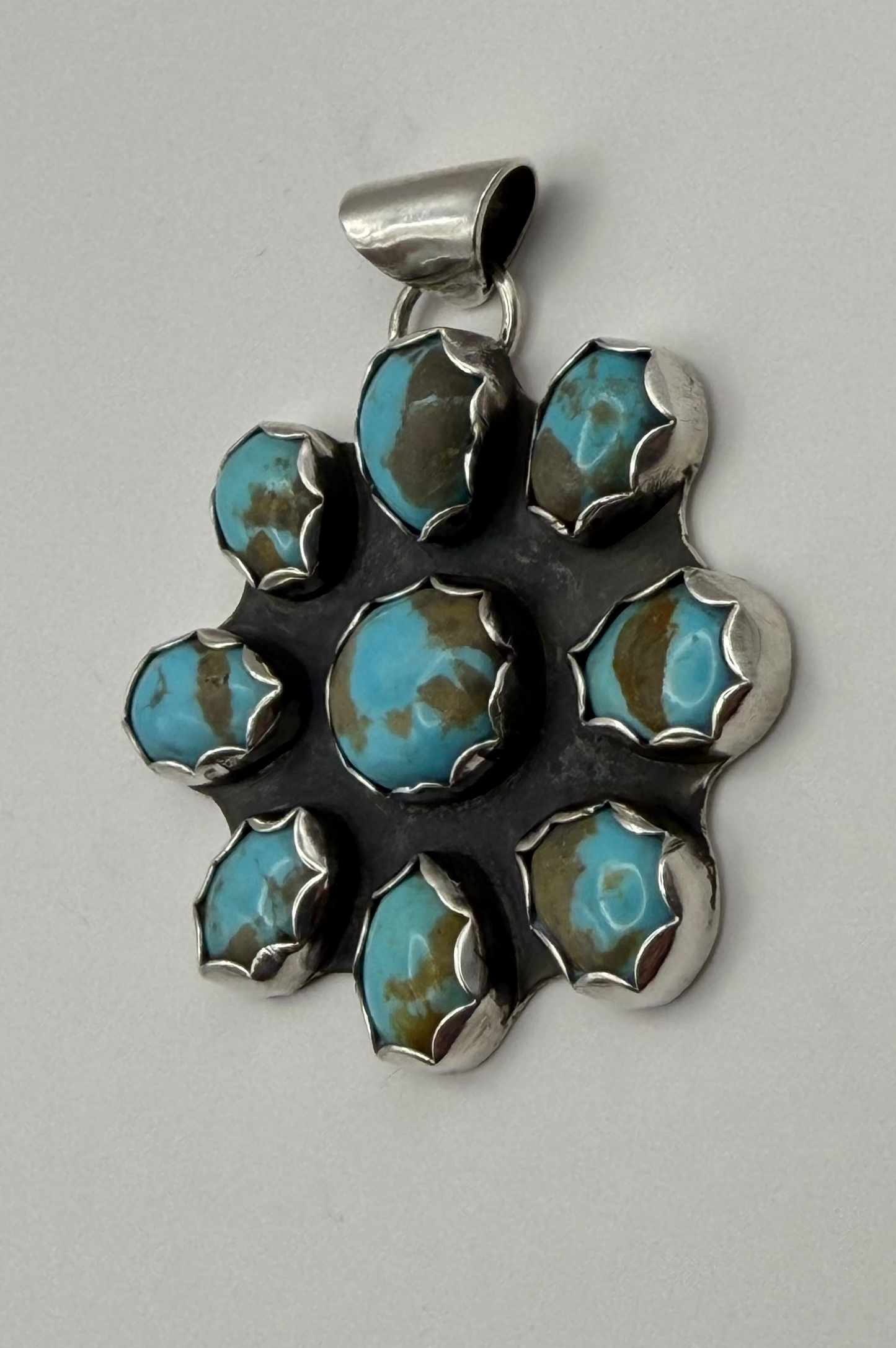 A sterling silver pendant featuring a cluster of seven oval turquoise stones with brown matrix, set in a floral or sunburst pattern on a white background.