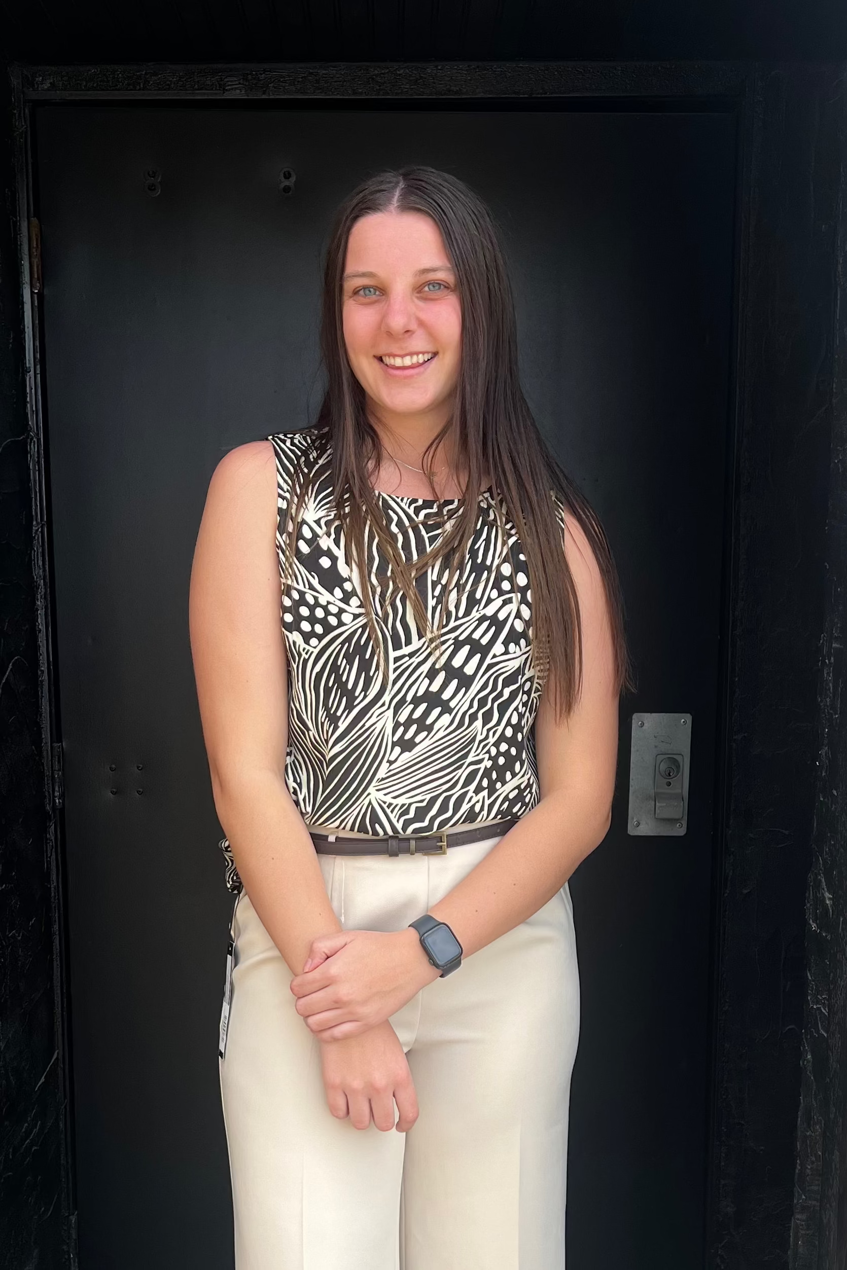 Woman standing in a dark doorway wearing a patterned top and white pants.