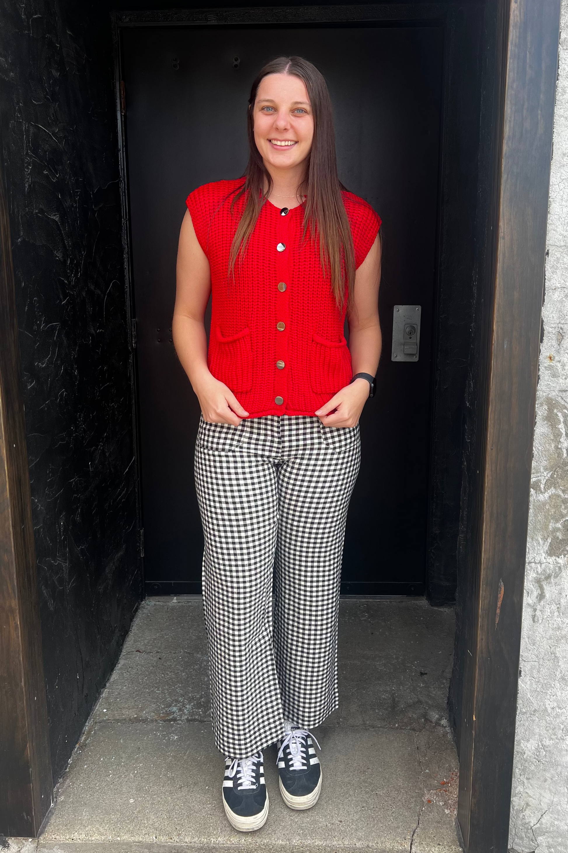 Person wearing a red sleeveless shirt, checkered pants, and sneakers standing in a doorway.