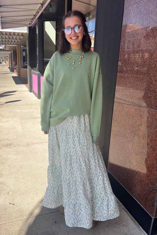 Woman in a green sweater and floral skirt standing on a sidewalk with a building in the background.