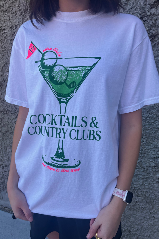 Person wearing a white t-shirt with a cocktail graphic and text against a gray wall.