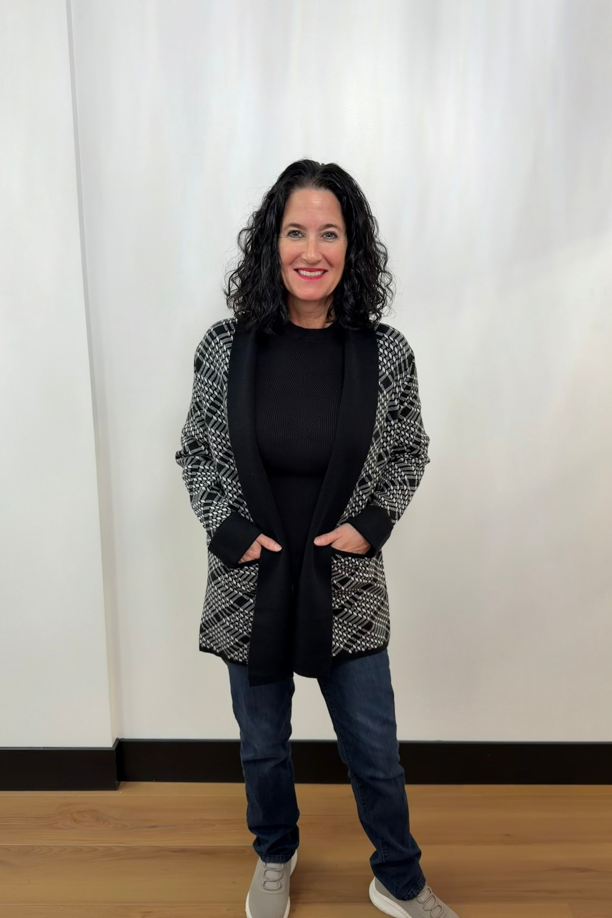 Woman wearing a black and white patterned cardigan over a black top and blue jeans, standing against a plain white background.