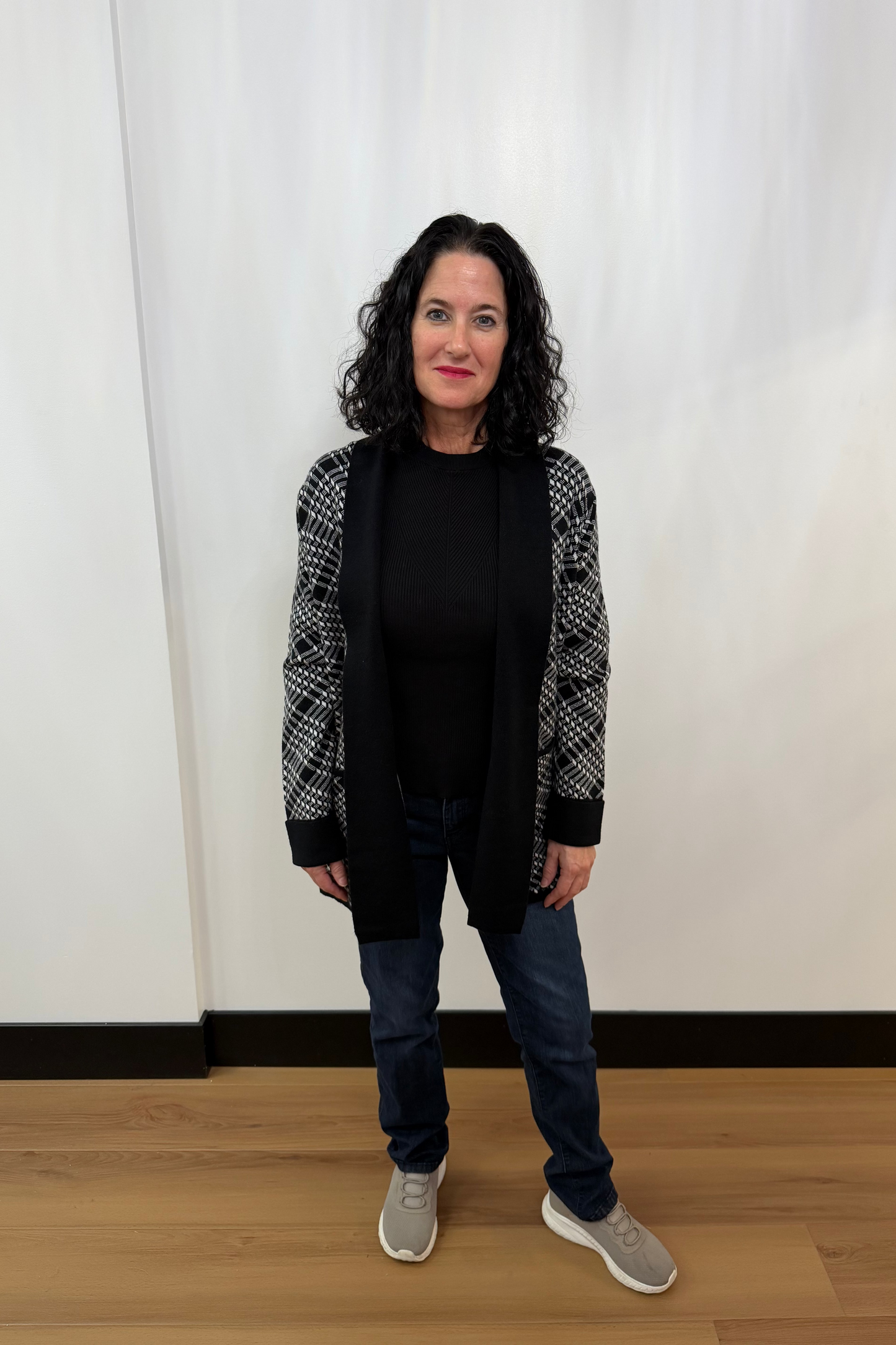 Woman wearing a black top, patterned cardigan, jeans, and gray sneakers against a white curtain background.