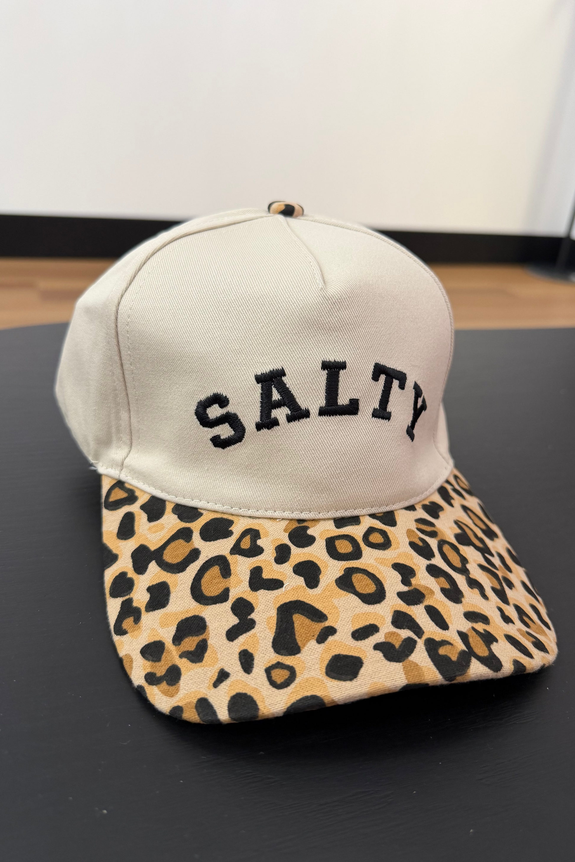 Beige cap with 'SALTY' text and leopard print brim on a black surface.