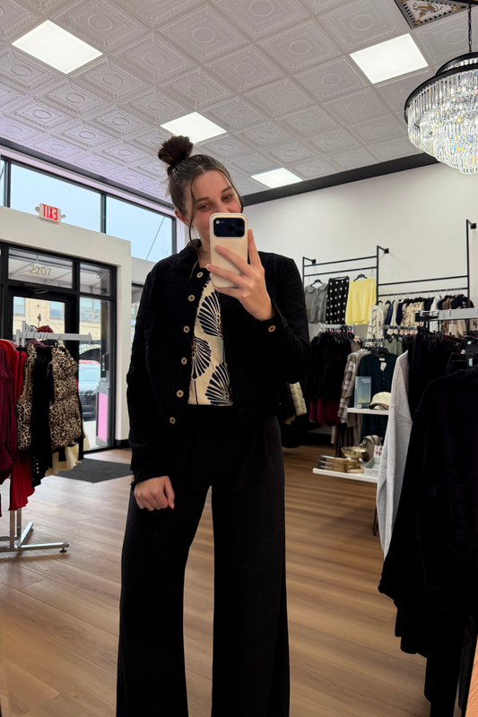 Person taking a mirror selfie in a clothing store wearing a black outfit with gold buttons.