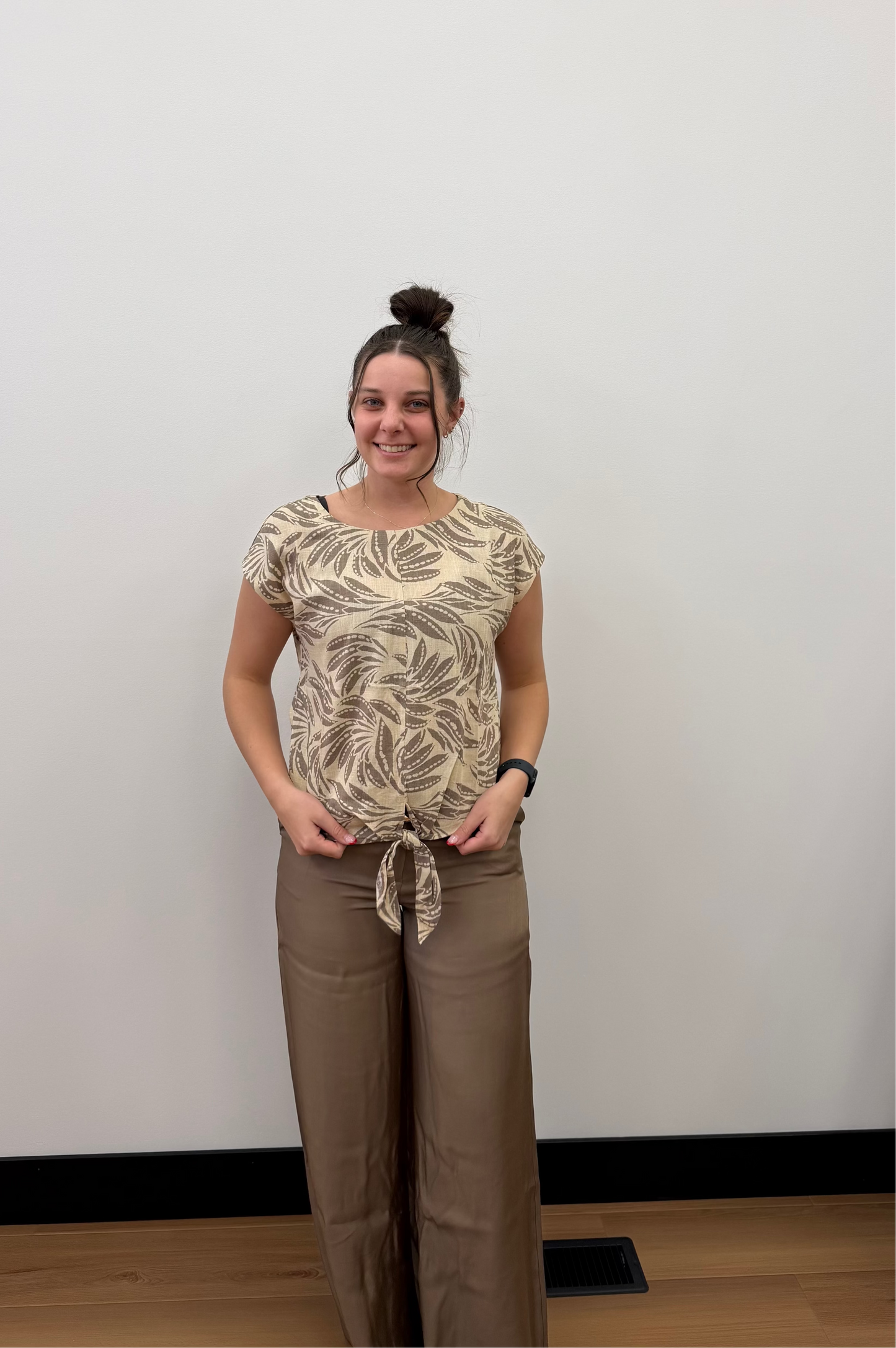 Woman wearing a patterned top and brown pants standing against a white wall.