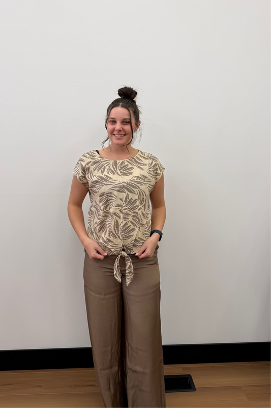 Woman wearing a patterned top and brown pants standing against a white wall.