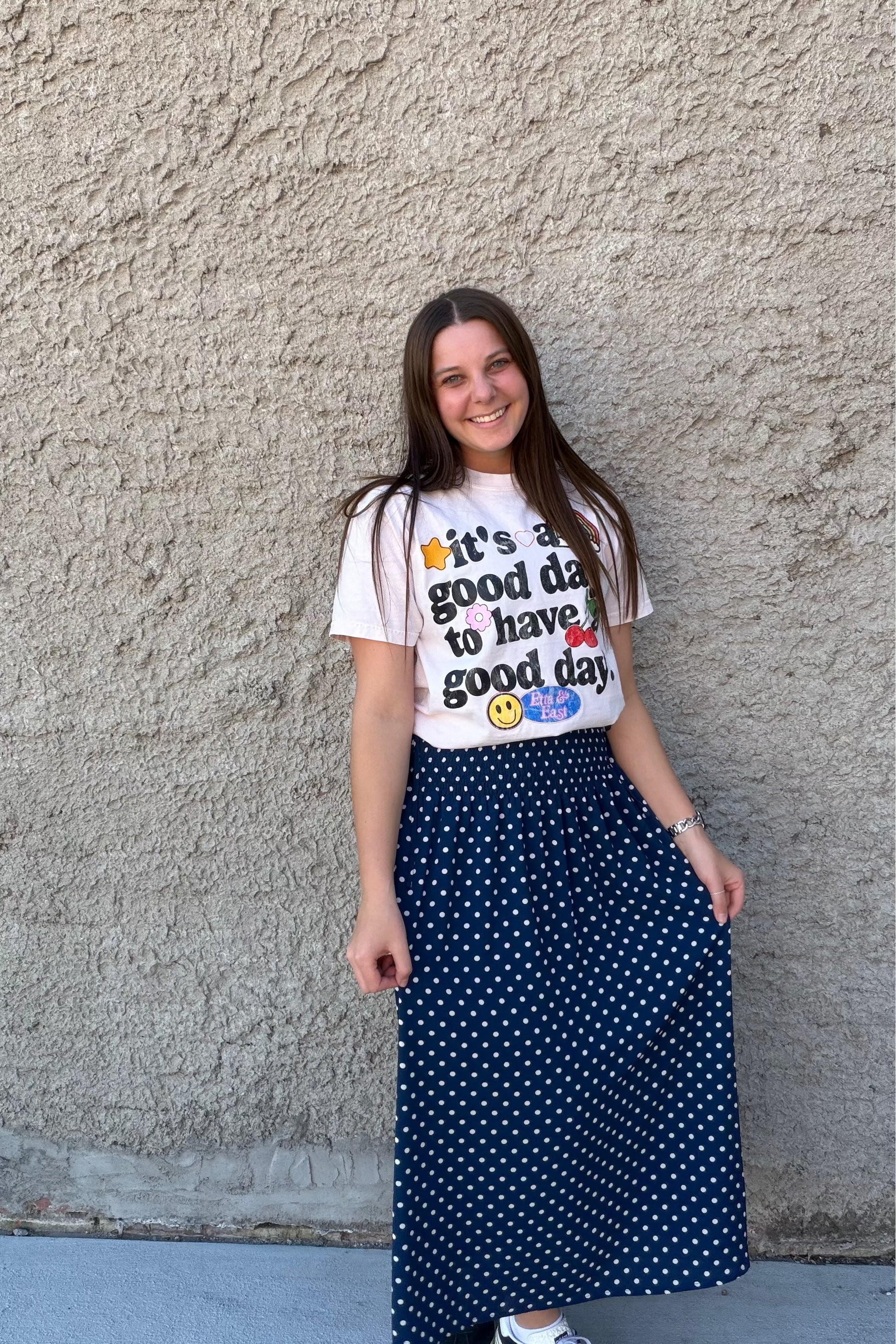 Woman wearing a white t-shirt with text and a blue polka dot skirt standing against a textured wall.