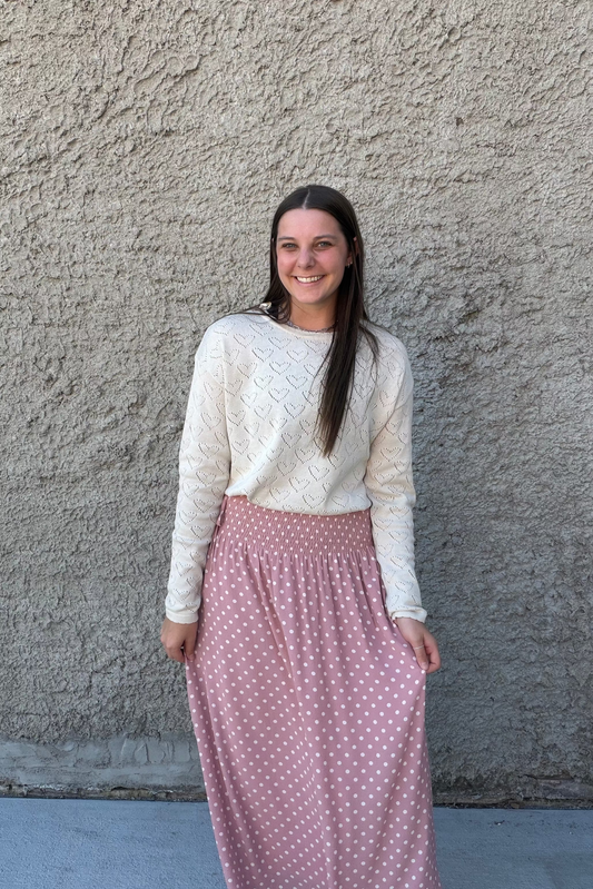 Woman wearing a white sweater and pink polka dot skirt standing against a textured wall.