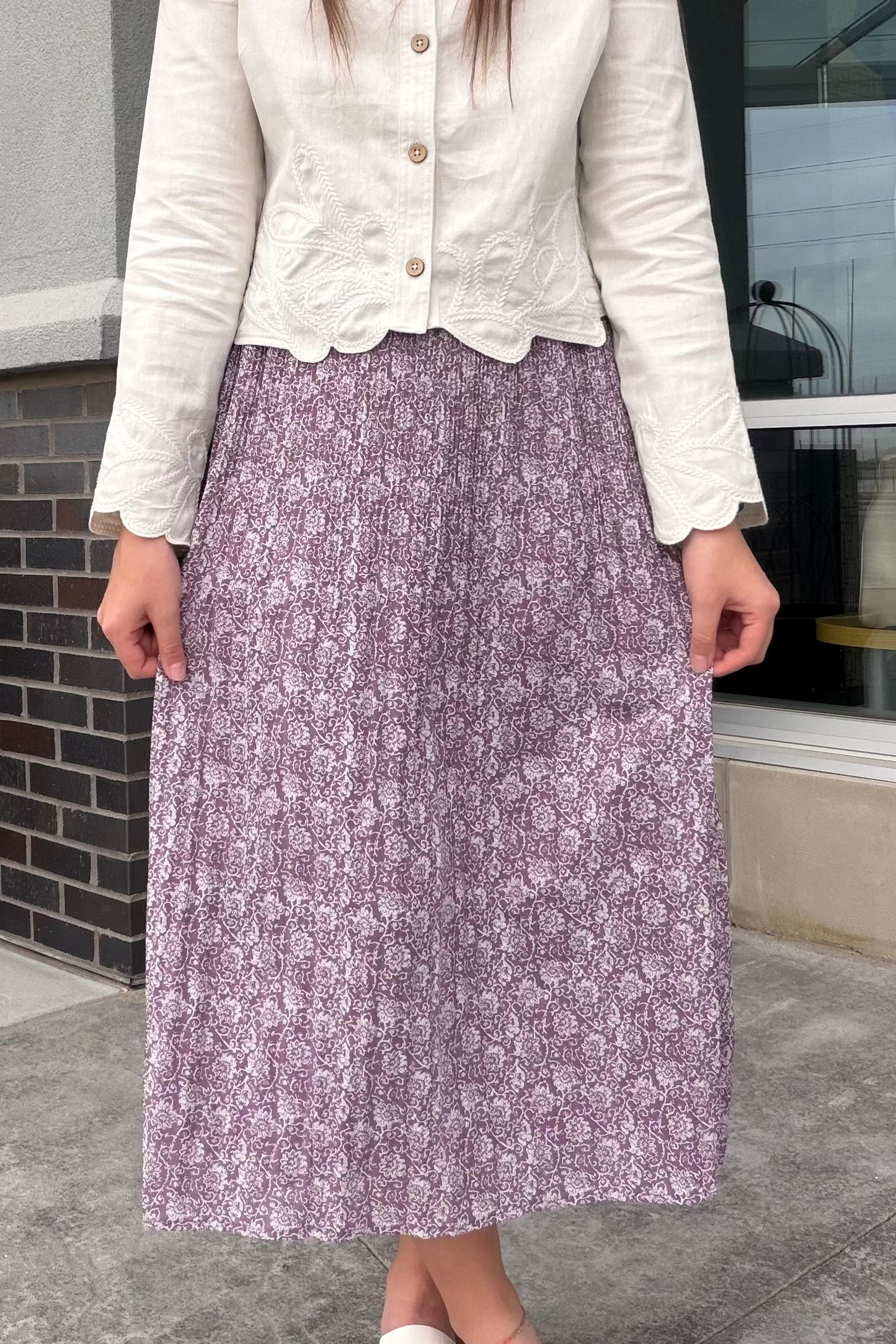 Person wearing a white blouse and purple floral skirt standing in front of a building.