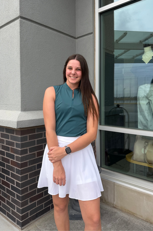 Woman in a teal sleeveless top and white skirt standing outside a building with a window display.