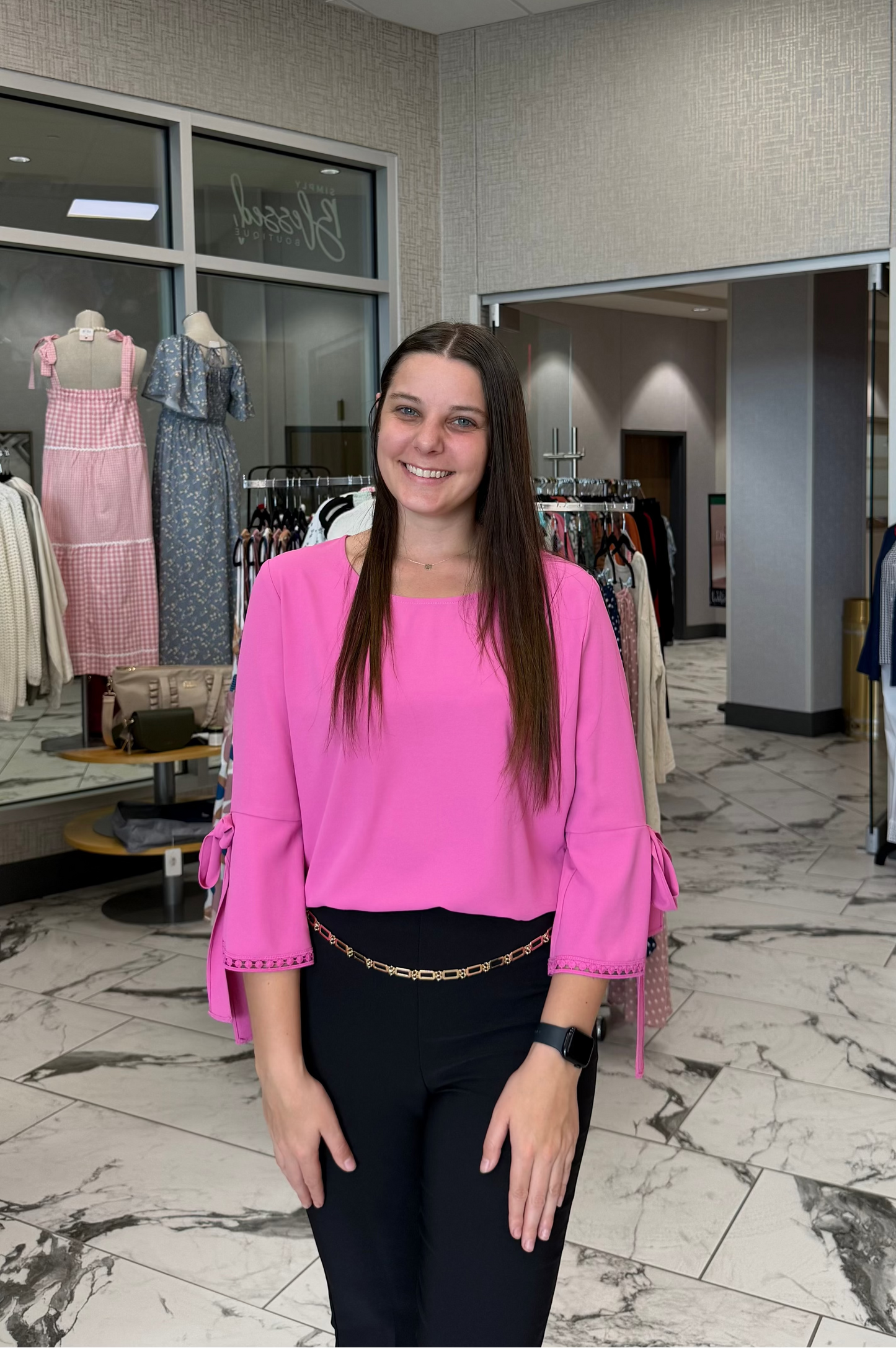 Woman in a pink top standing in a clothing store with racks of clothes in the background.