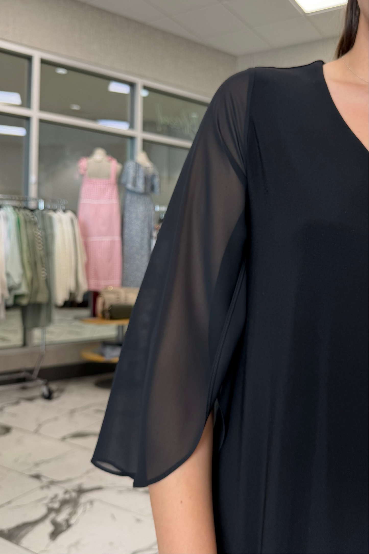 Person wearing a black outfit in a clothing store with mannequins and dresses in the background.