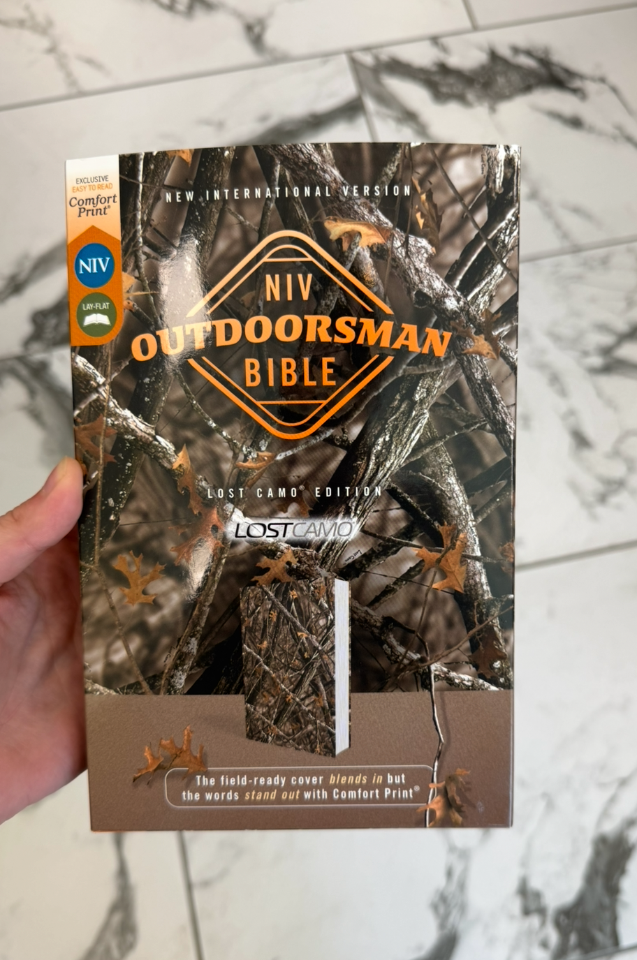 Hand holding an 'HIV Outdoorsman Bible' on a marble surface