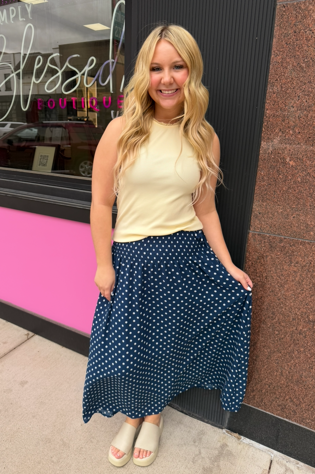 Woman in a yellow top and blue polka dot skirt standing in front of a store with 'Blessed' sign.