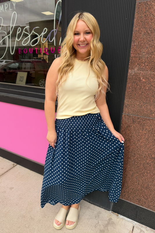 Woman in a yellow top and blue polka dot skirt standing in front of a store with 'Blessed' sign.