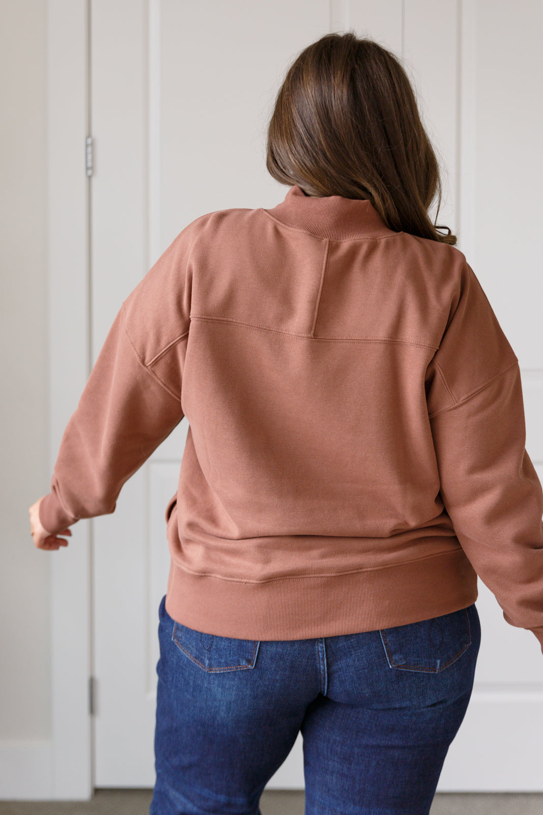 Person wearing a brown sweater and blue jeans against a white wall