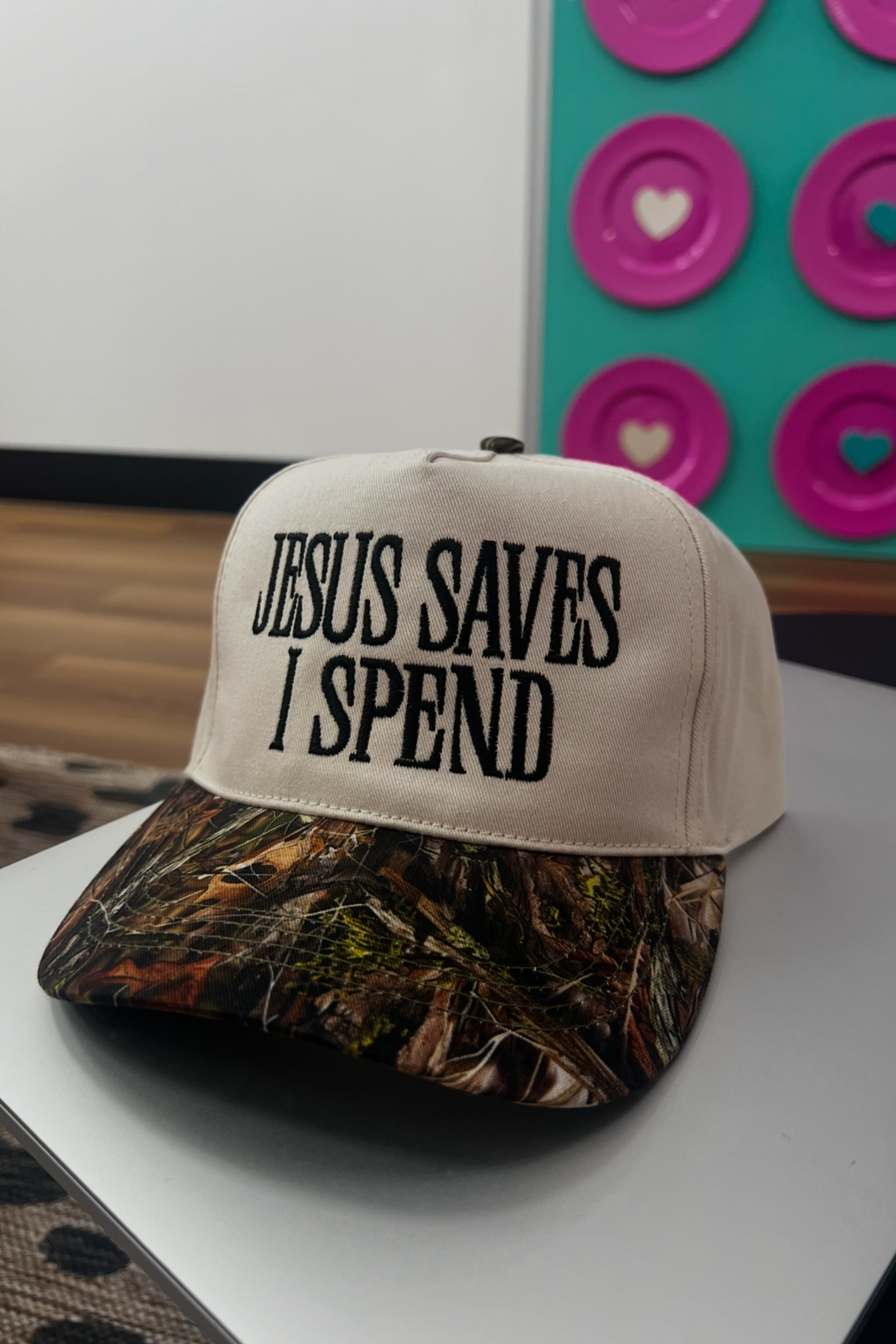 Beige cap with 'Jesus Saves I Spend' text on a white surface with colorful plates in the background.