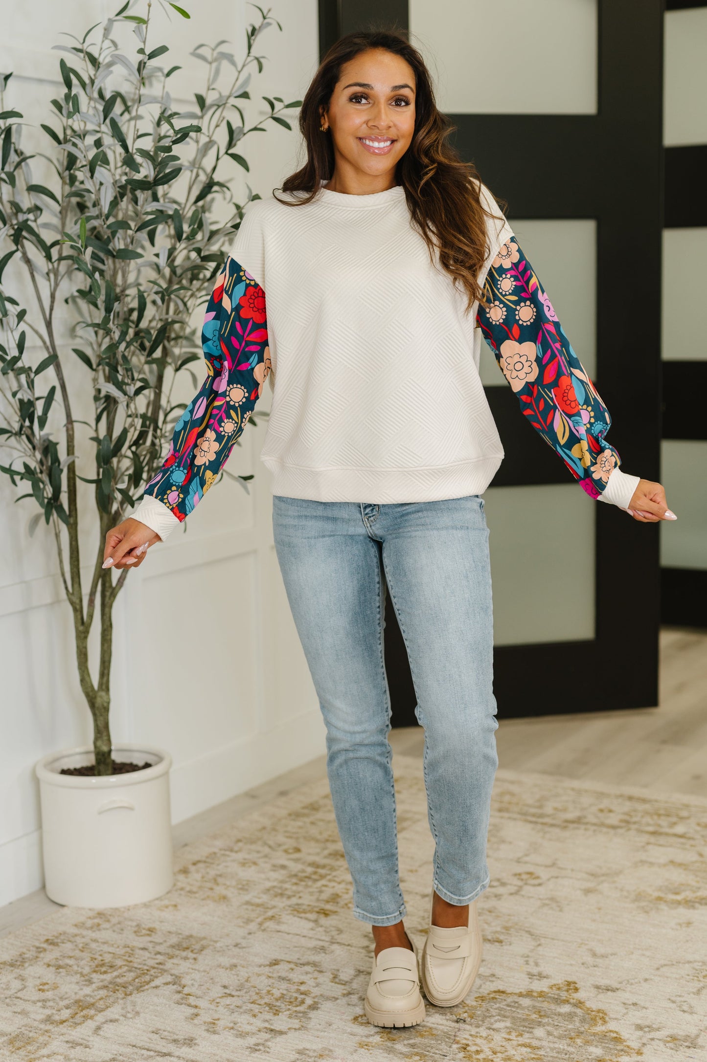 Woman wearing a white sweater with colorful sleeves and light blue jeans, standing indoors.