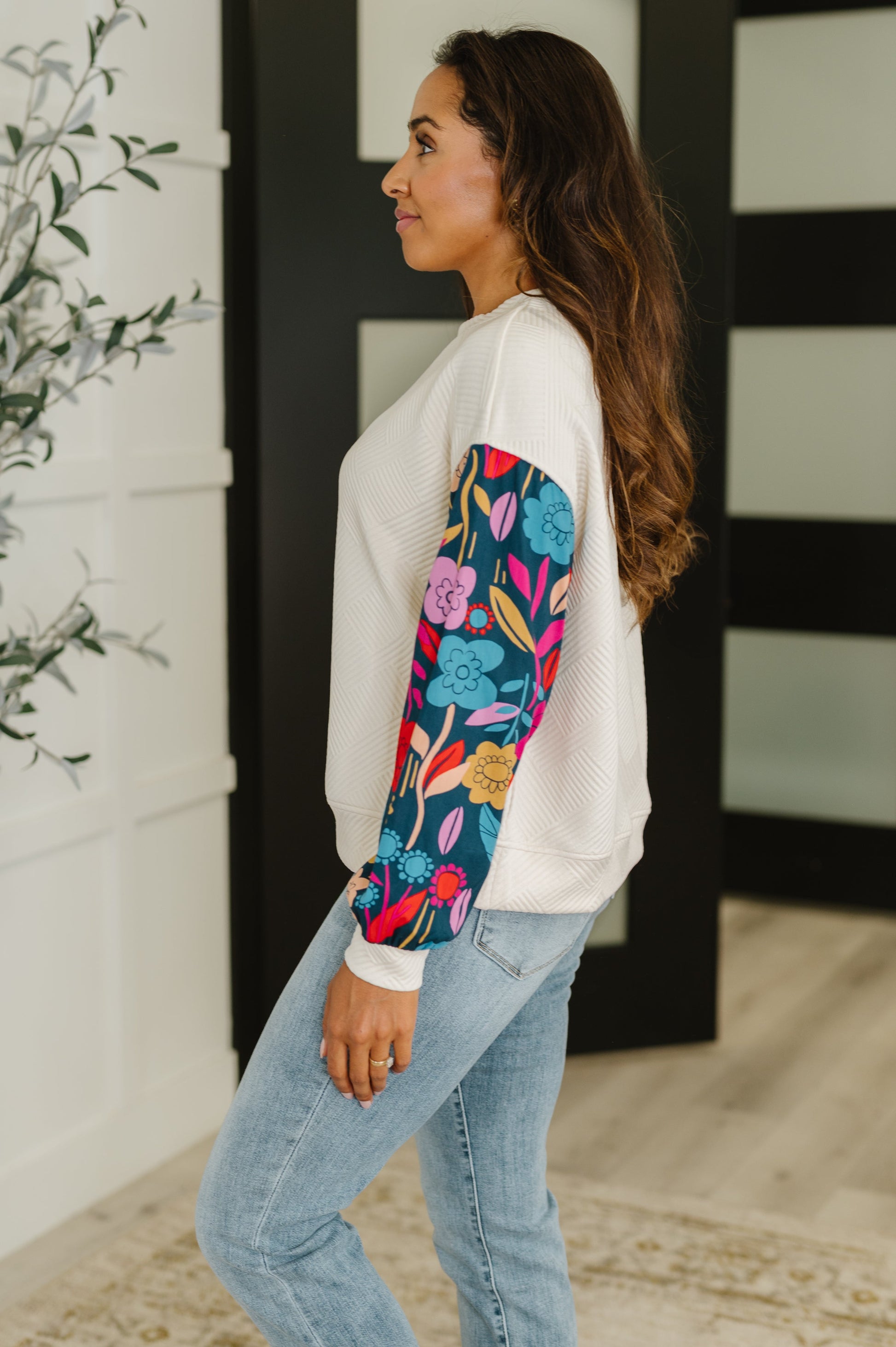 Woman wearing a white sweatshirt with colorful floral sleeves and blue jeans indoors.