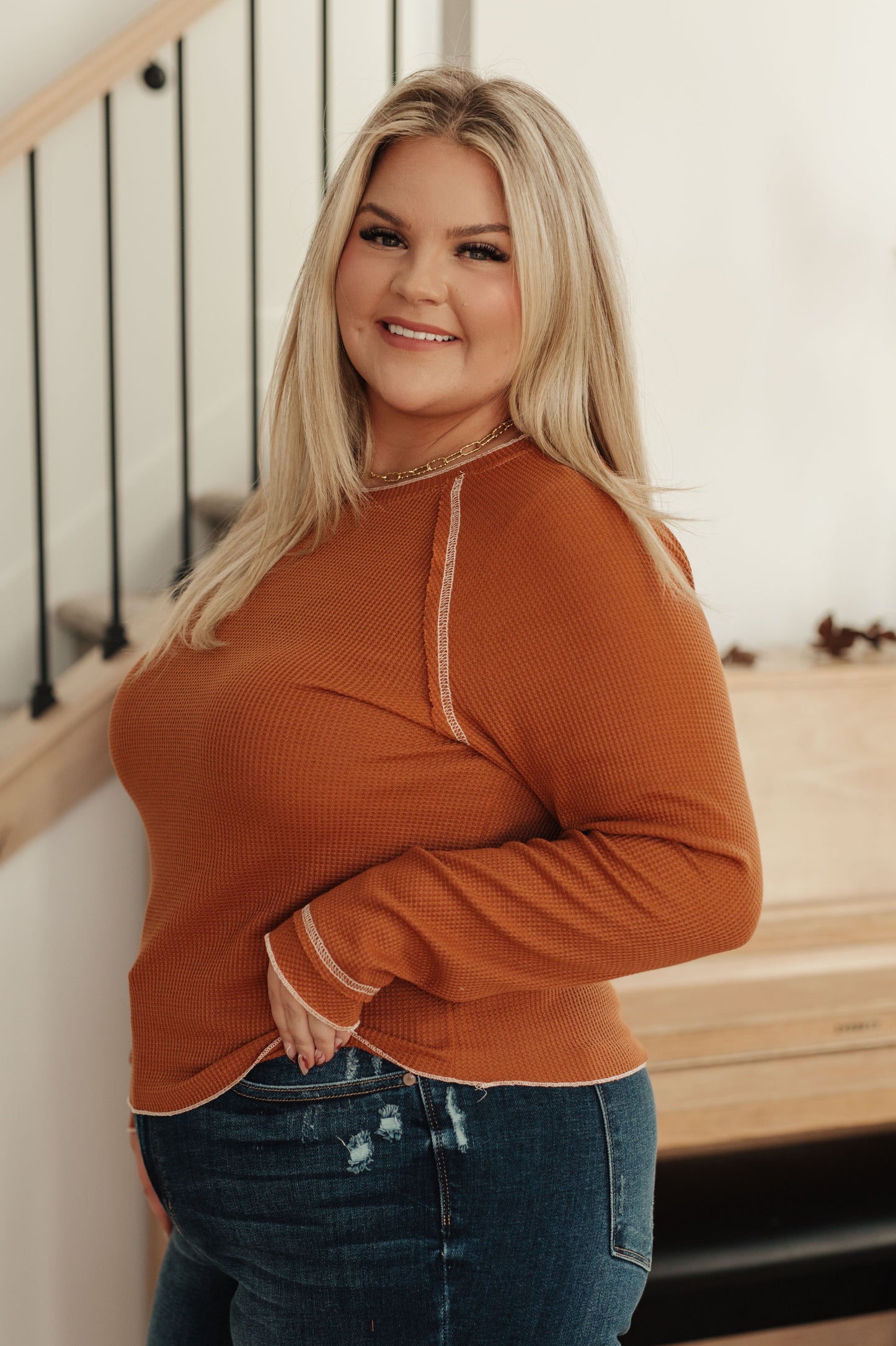 Woman wearing a rust long-sleeve top and blue jeans indoors.