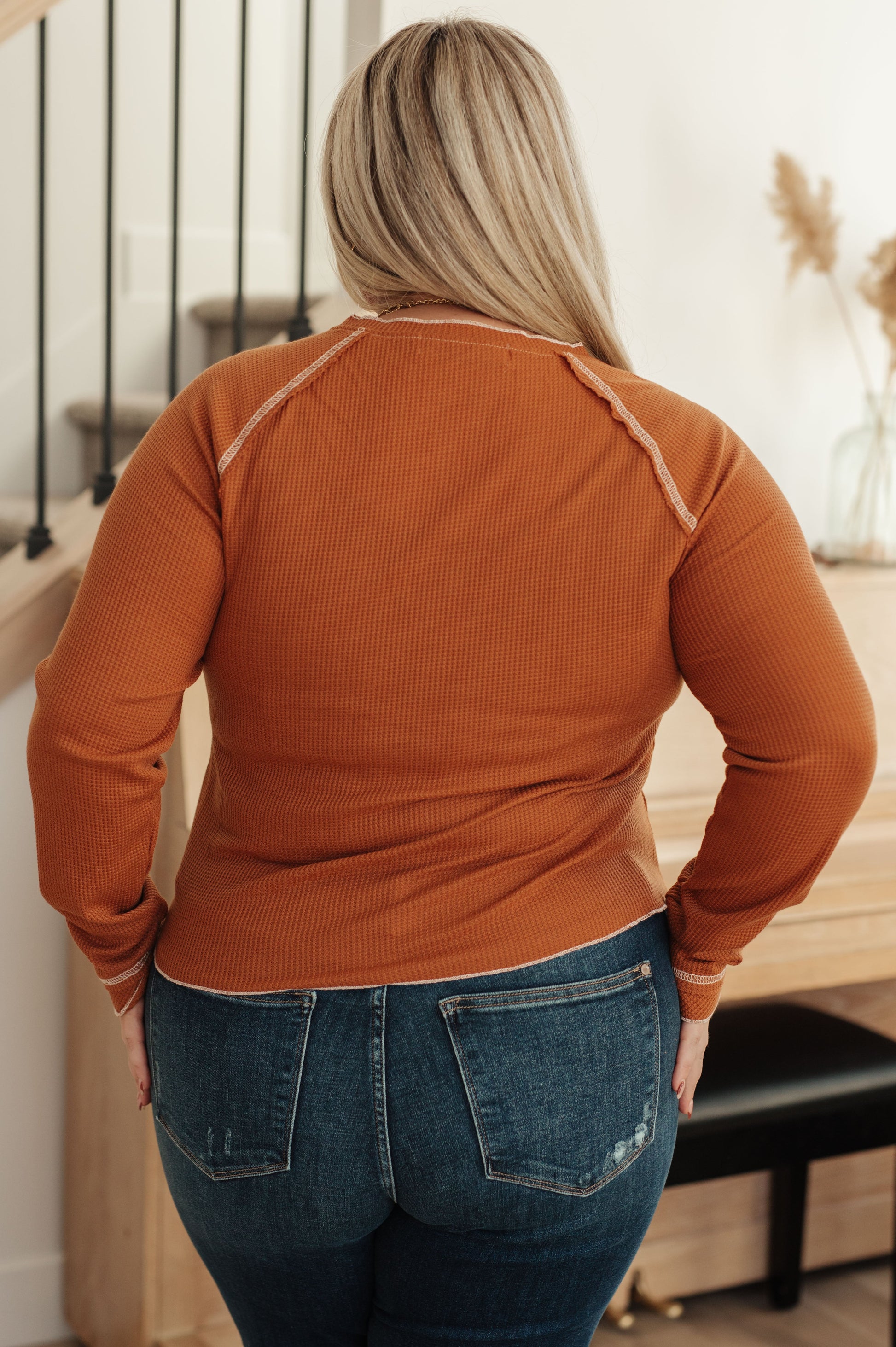 Woman wearing a rust long-sleeve top and blue jeans indoors.