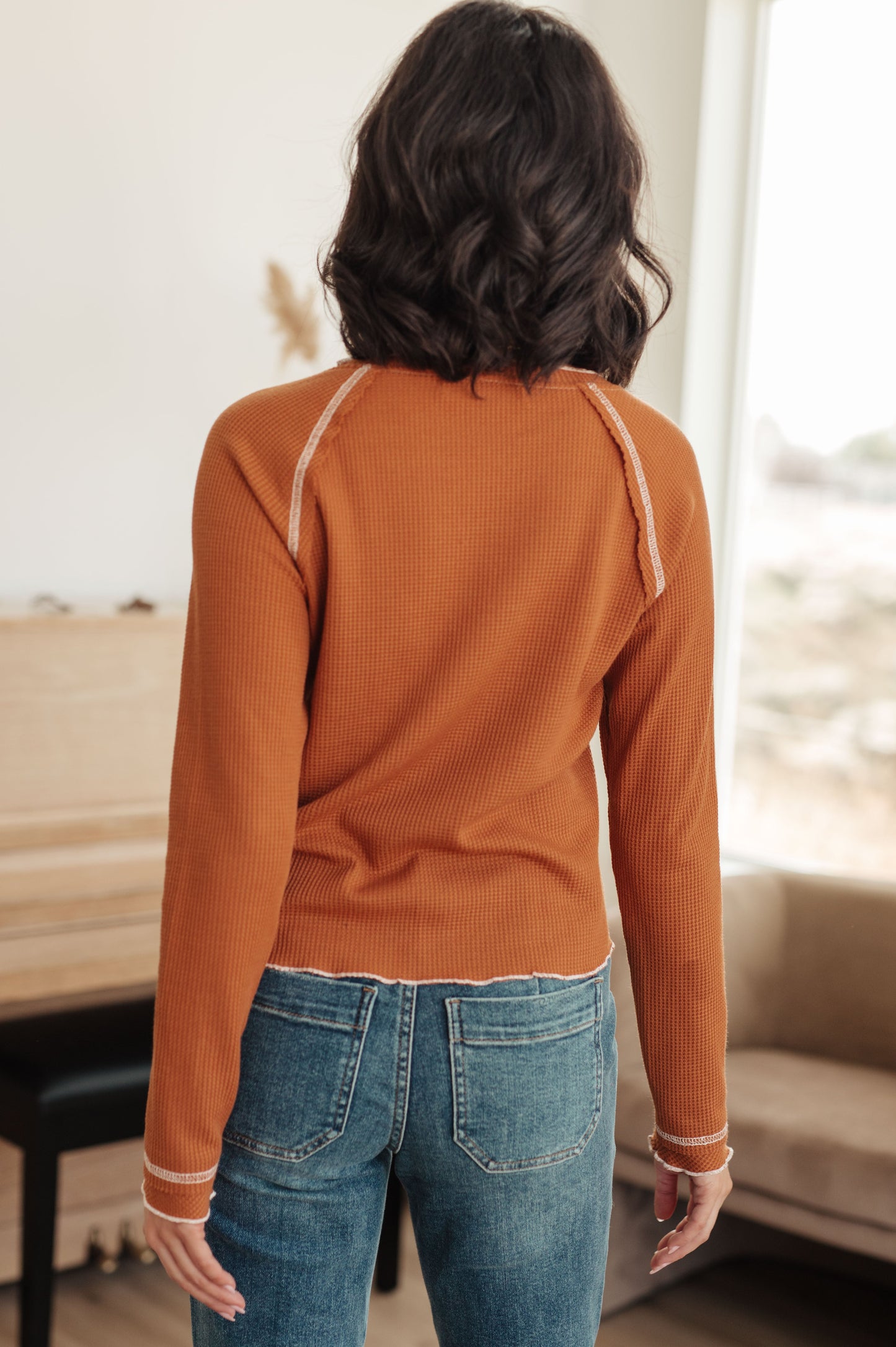 Woman wearing a rust long-sleeve top and blue jeans indoors.