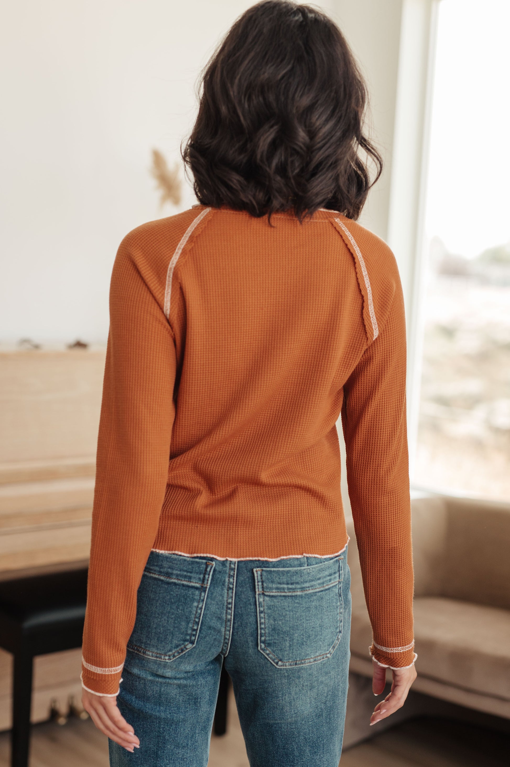 Woman wearing a rust long-sleeve top and blue jeans indoors.