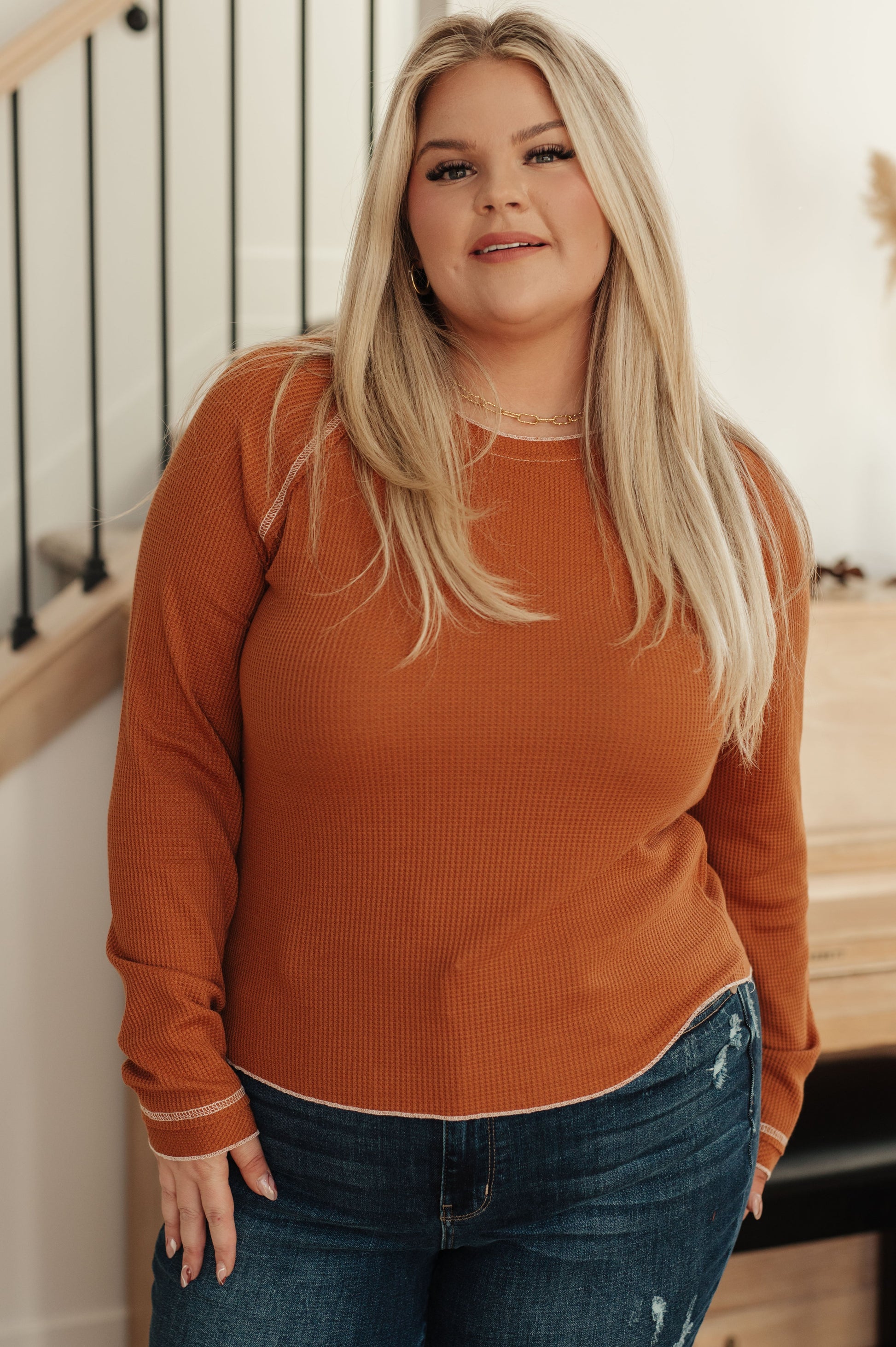 Woman wearing a rust long-sleeve top and blue jeans indoors.