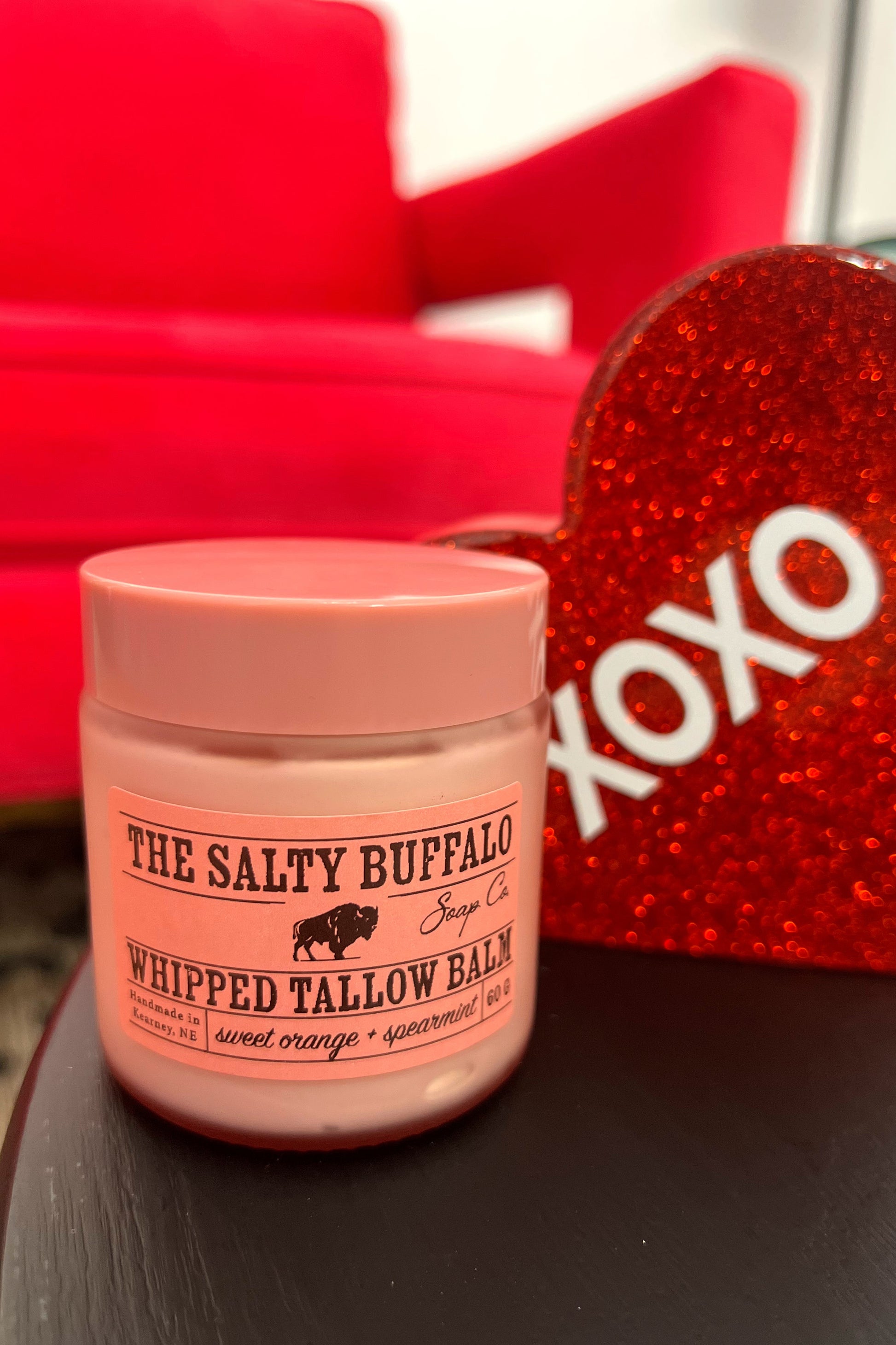 Pink jar of 'The Salty Buffalo' whipped tallow balm on a dark surface with a red heart-shaped object in the background.