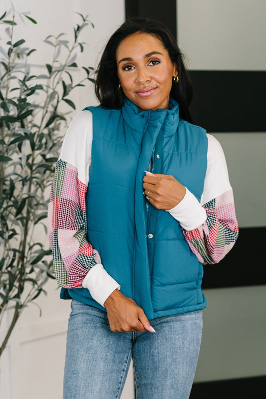 Woman wearing a teal puffer vest over a colorful plaid shirt with a blurred indoor background