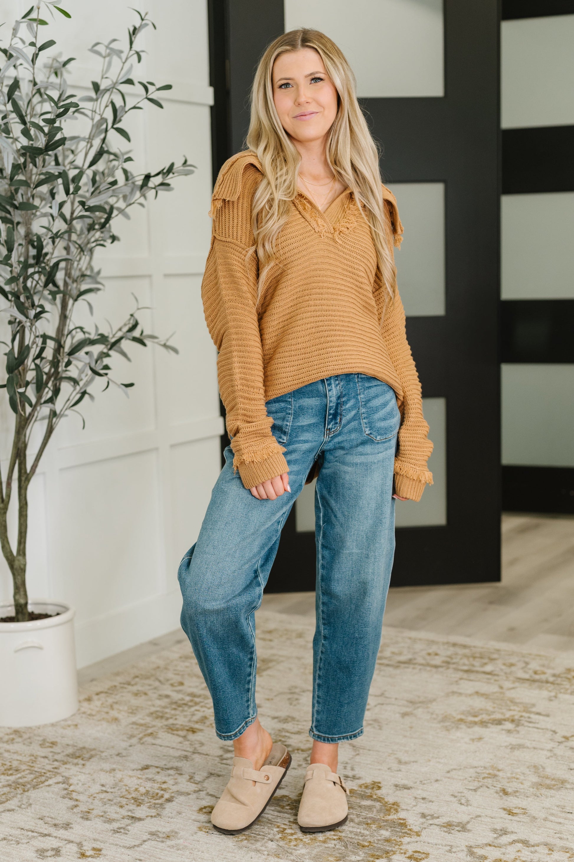 Woman wearing a brown sweater and blue jeans standing indoors with a plant in the background.