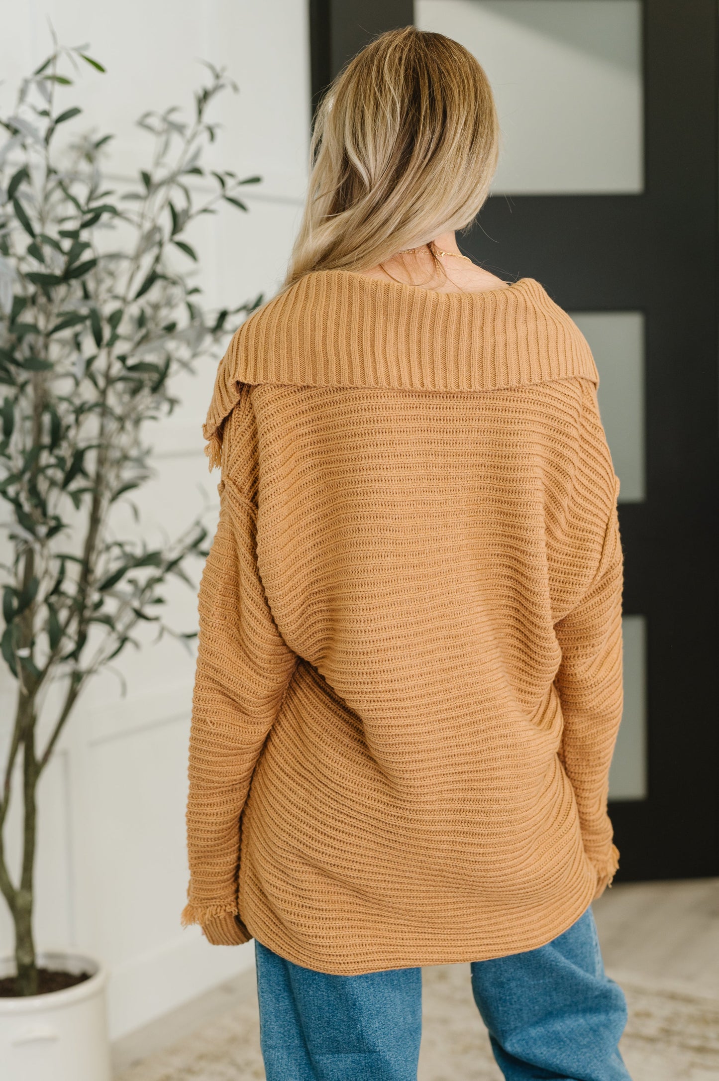 Person wearing a brown sweater in an indoor setting with a plant and door in the background