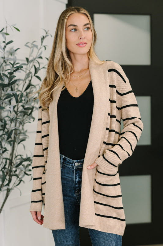 Woman wearing a beige cardigan with black stripes, black top, and blue jeans indoors.