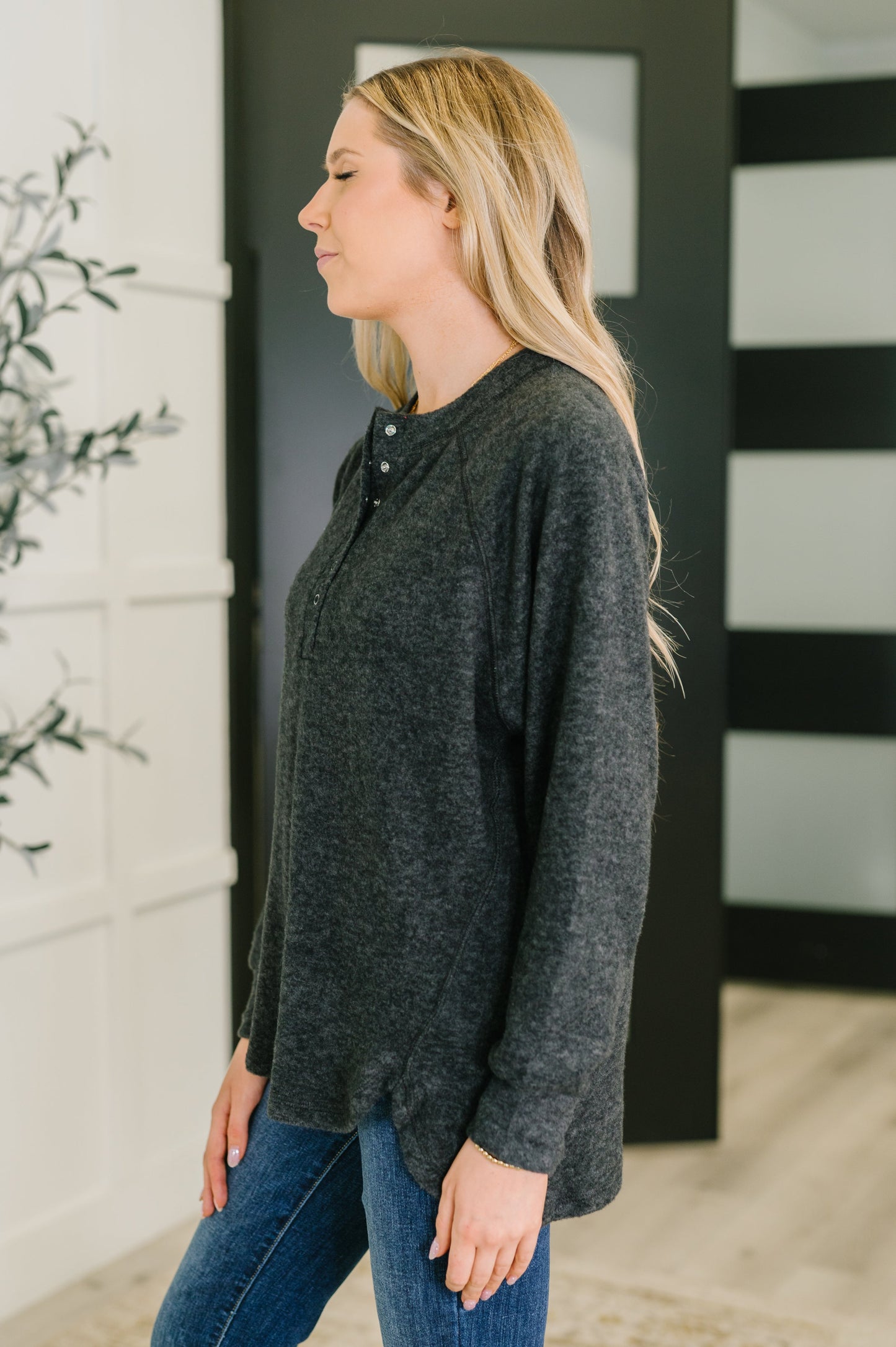 Woman wearing a dark gray long-sleeve top and blue jeans standing against a black and white striped wall.