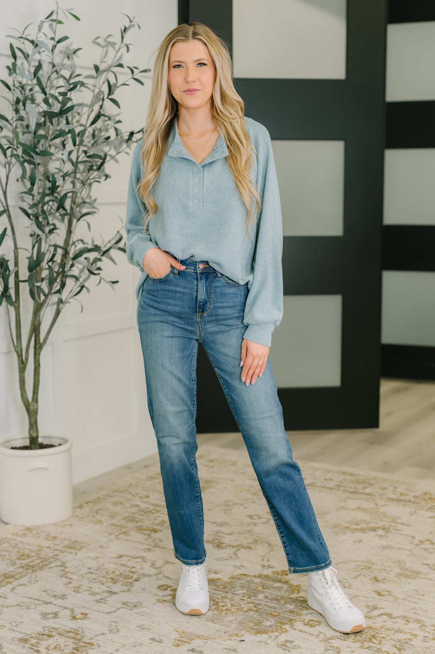 Woman wearing a light blue denim shirt and jeans standing indoors with a plant and decorative wall in the background.