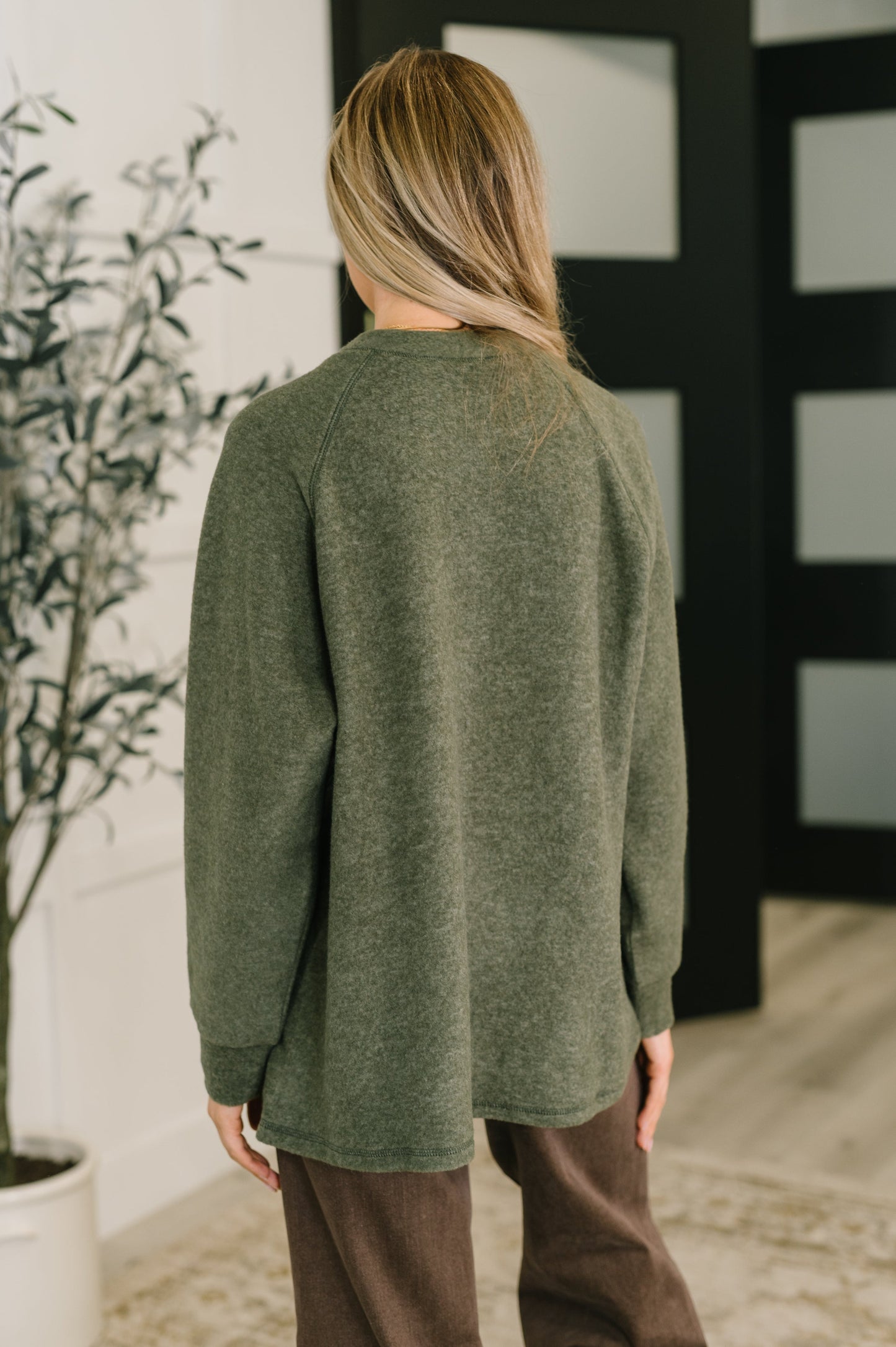Person wearing a green sweater and brown pants standing indoors with a plant and wall decor in the background.