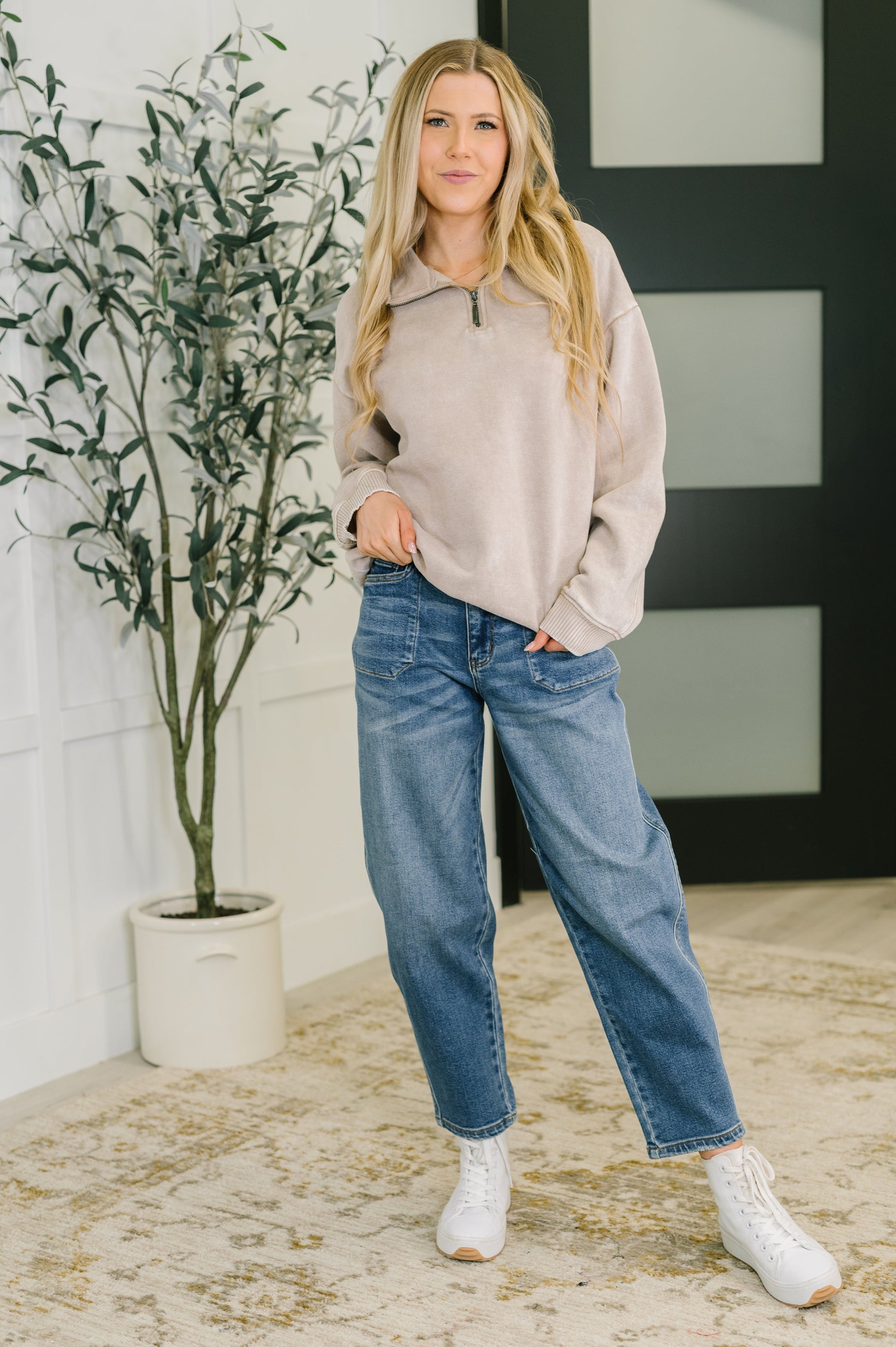 Woman wearing a beige sweater and blue jeans standing indoors with a plant in the background