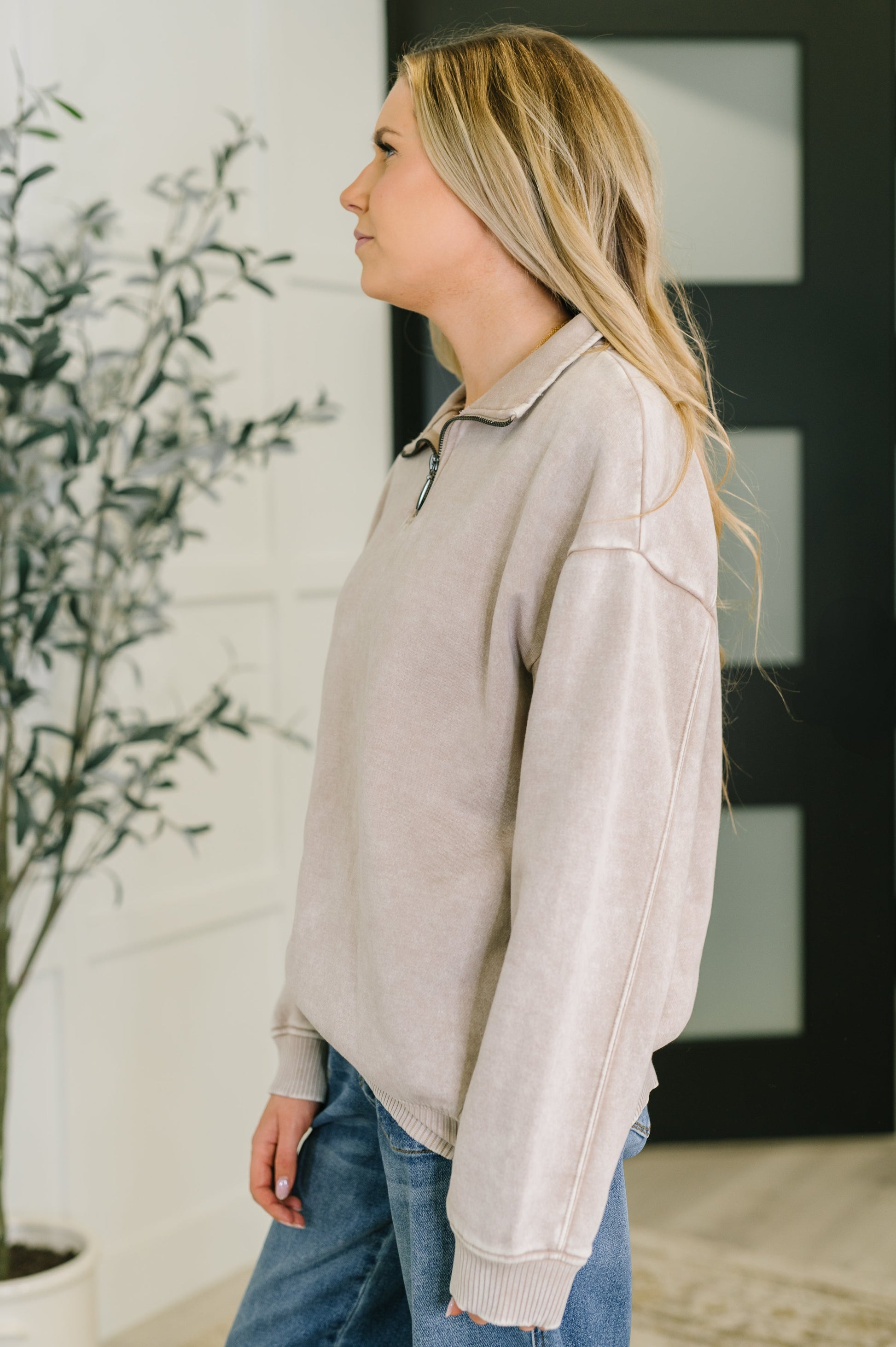 Woman wearing a beige sweater and blue jeans standing indoors next to a plant.