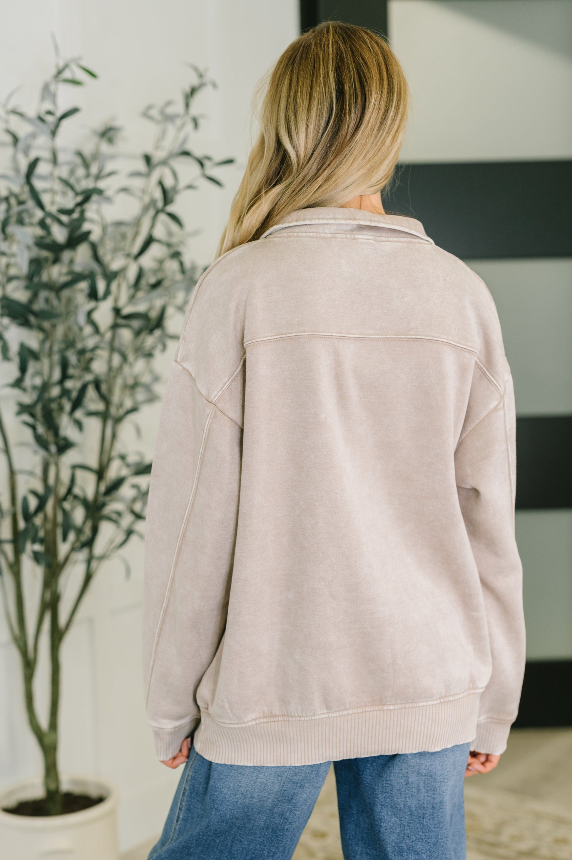 Person wearing a beige jacket and blue jeans standing in a room with a plant.
