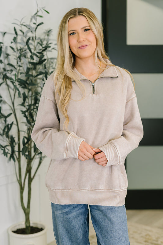 Woman wearing a beige pullover with a zipper, standing indoors next to a plant.