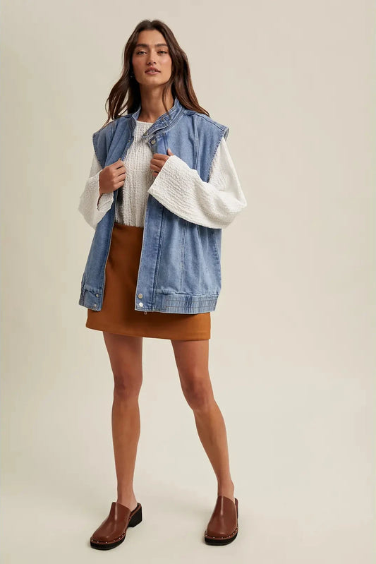 Woman wearing a denim vest over a white sweater and brown skirt on a beige background