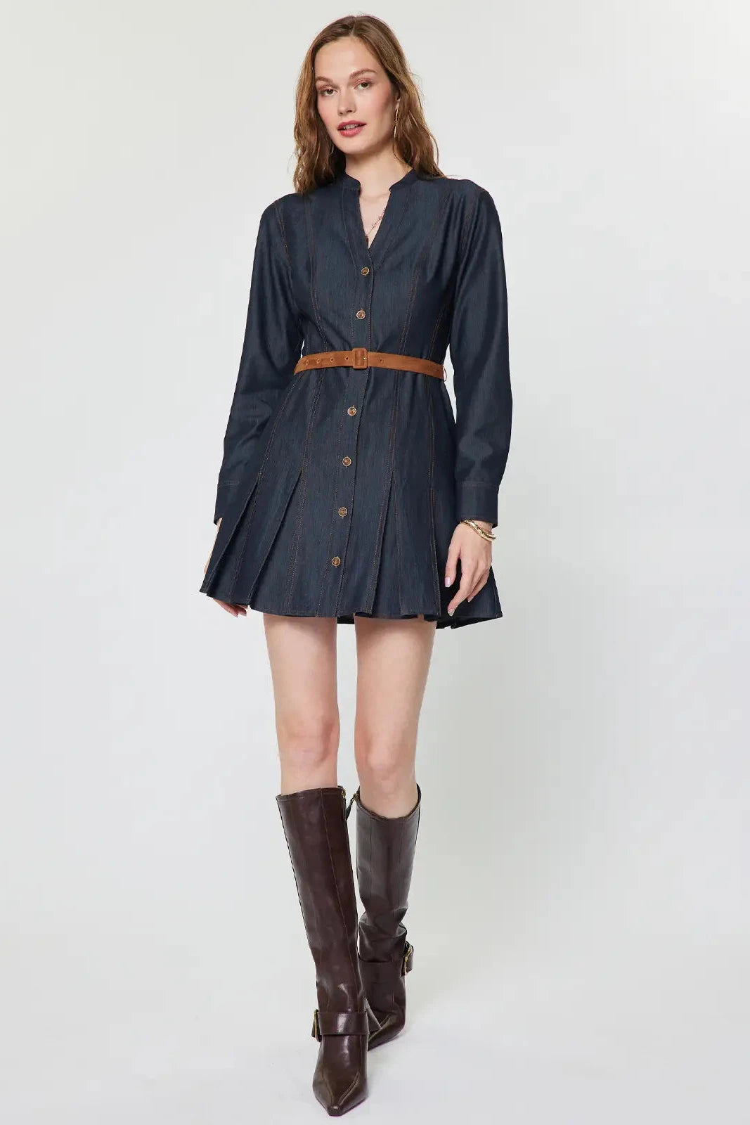 A woman models a dark navy blue denim shirt dress with a brown belt and brown knee-high boots, standing against a light gray studio background.