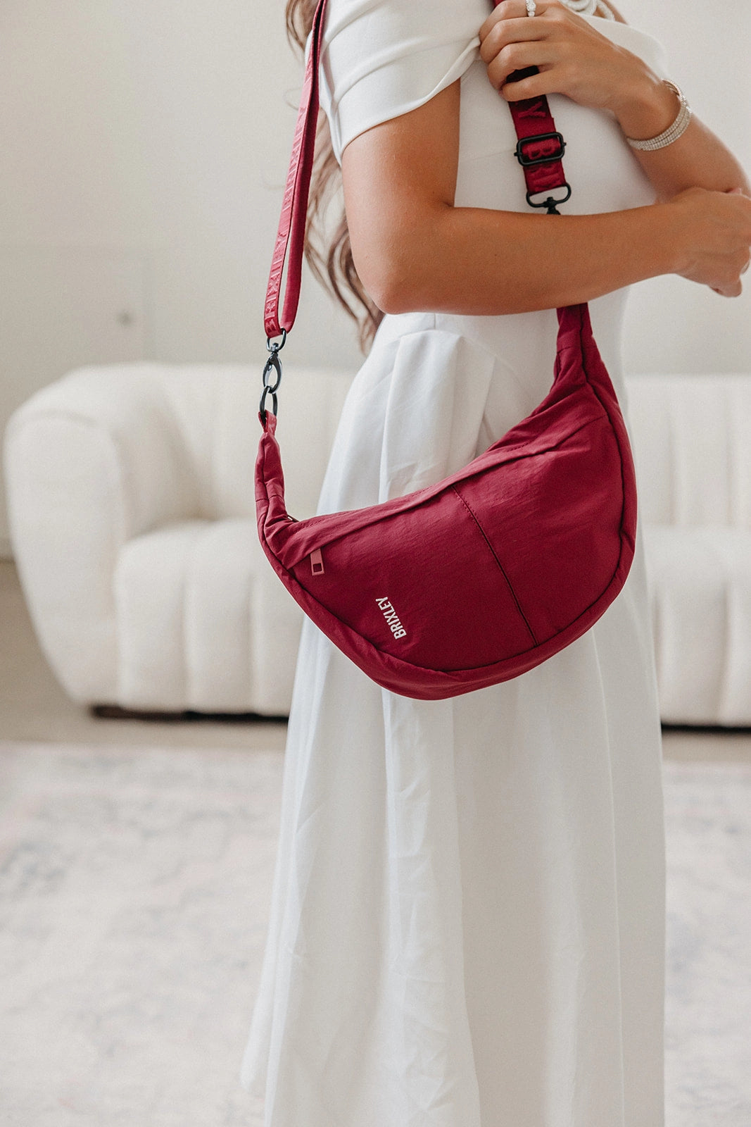 A woman in a white dress wearing a red crescent-shaped sling bag with a matching shoulder strap in a bright, modern room.